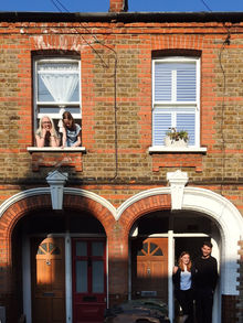 Two caucasian people, a mum and a daughter lean out of a top floor window and down stairs a couple stand in their doorway of a red brick house.  It is a doorstep portrait photograph taken during the coronavirus pandemic lockdown in London.  It is a coronavirus lockdown covid-19 doorstep portrait from empowerment photographer HeardinLondon.