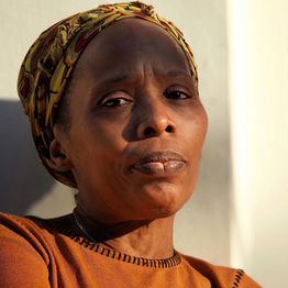 A black woman in an orange top and a headscarf looks at the camera.  It is a doorstep portrait photograph taken during the coronavirus pandemic lockdown in London.  It is a coronavirus lockdown covid-19 doorstep portrait from empowerment photographer HeardinLondon.