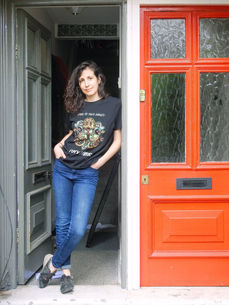 An Irish Italian woman stands leaning into her doorway, there is a red door to her right. It is a doorstep portrait photograph taken during the coronavirus pandemic lockdown in London.  It is a coronavirus lockdown covid-19 doorstep portrait from empowerment photographer HeardinLondon.