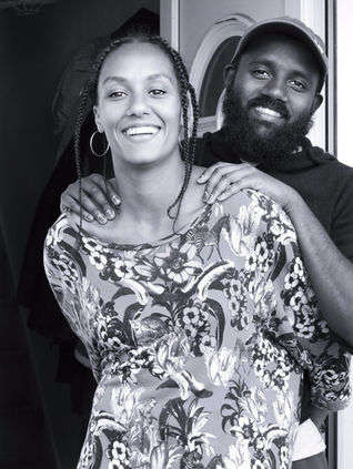 A black and white portrait photograph of a black couple in love, she is at the front with plaits and a patterned dress and he is stood behind her wearing a cap, both are smiling at the camera.  It is a coronavirus lockdown covid-19 doorstep portrait from empowerment photographer HeardinLondon.