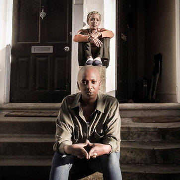 A St Lucian mother and daughter sit on the step outside their home, the mother is at the top and the daughter sits closer to the camera.  Both are looking into the lens. It is a doorstep portrait photograph taken during the coronavirus pandemic lockdown in London.  It is a coronavirus lockdown covid-19 doorstep portrait from empowerment photographer HeardinLondon.