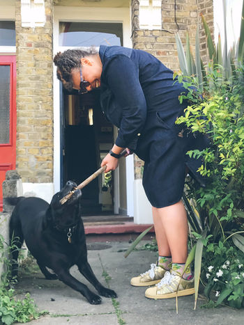 A black woman in a blue dress plays with her black dog and it bites a stick in her hand.  It is a doorstep portrait photograph taken during the coronavirus pandemic lockdown in London.  It is a coronavirus lockdown covid-19 doorstep portrait from empowerment photographer HeardinLondon.