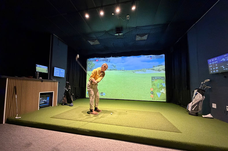 A female golfer wearing jacket and pants swings her golf club in front of an indoor golf simulator screen.