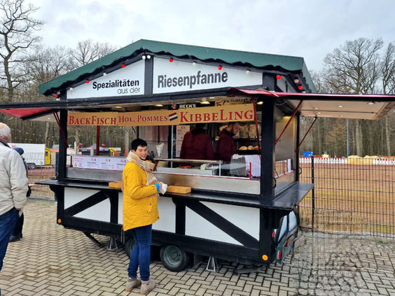 Frau am Imbissstand mit "Spezialitäten", "Riesenpfanne", "Backfisch", "Pommes", "Kibbeling"