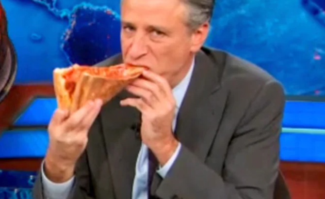 A picture of Jon Stewart eating a slice of pizza.