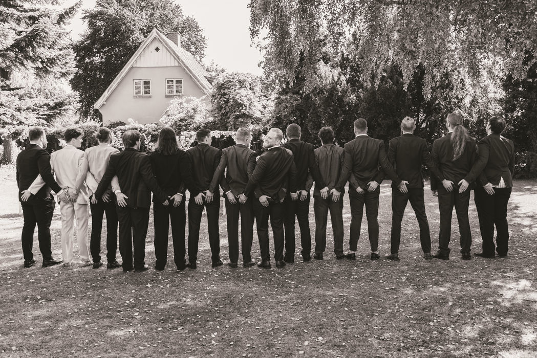 Gommens venner står på række til et sjovt bryllupsgruppebillede udendørs (sort/hvid)