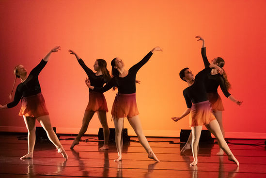 Five dancers with one arm each extended on an orange lit stage