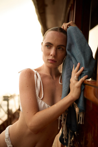 Woman in white bikini leaning on wood, holding a blue towel, Riana Yacht
