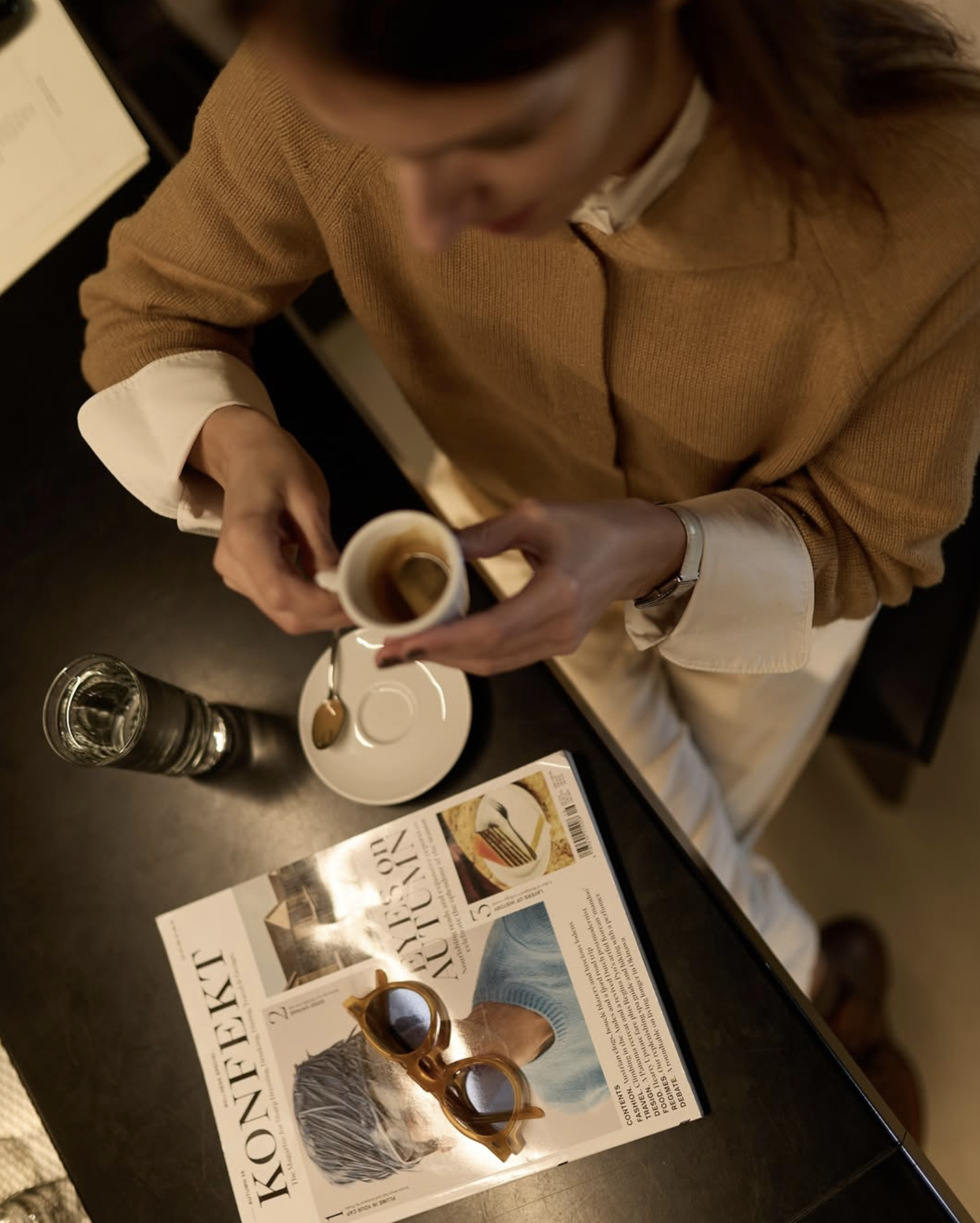 Woman drinking coffee, magazine KONFEKT, sunglasses, seated at table, top view.