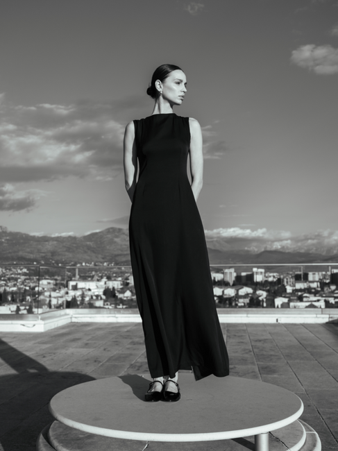 Woman in black dress poses, city and mountains in background on rooftop.