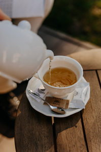 Pouring tea into a white cup, tea, sugar, and spoon on a wooden table.