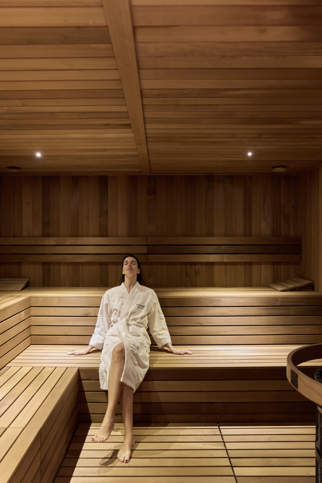 Woman relaxing in a wooden sauna, arms outstretched, dressed in bathrobe.