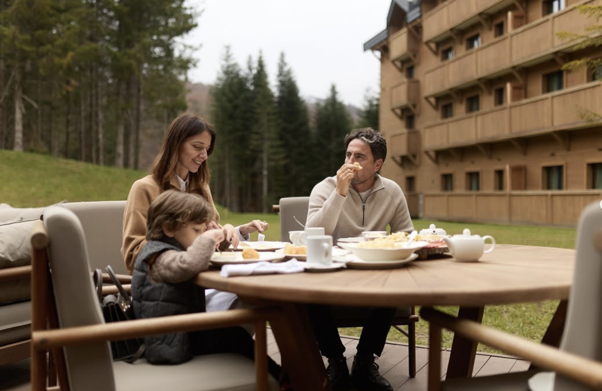Family eating breakfast outdoors, smiling, near a wooden building and mountains