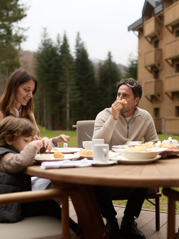 Family eating breakfast outdoors, smiling, near a wooden building and mountains