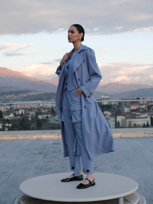 Woman wearing blue suit and coat, posing on rooftop, city background with mountains.