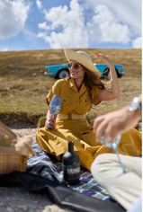 Woman in yellow dress holds glass, blue car in background, picnic setting.