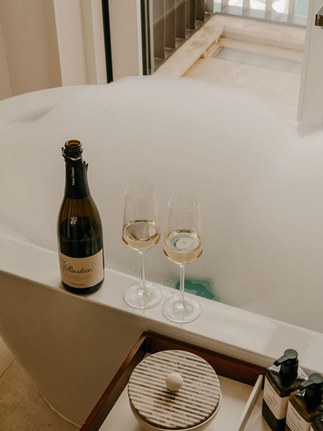Bottle of wine and two glasses next to a bubble-filled bathtub.