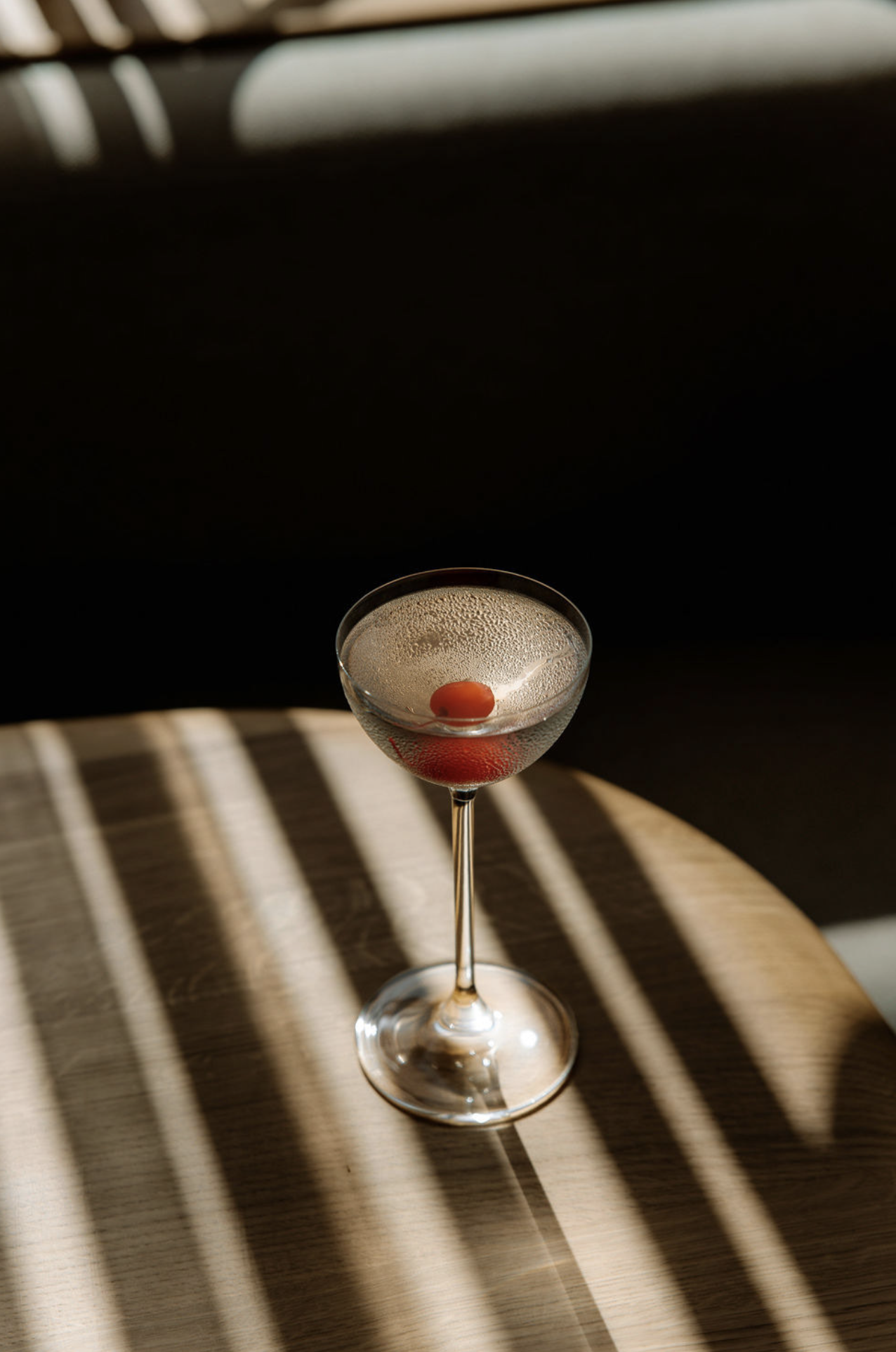 Elegant cocktail with cherry garnish on a table with striped shadows.