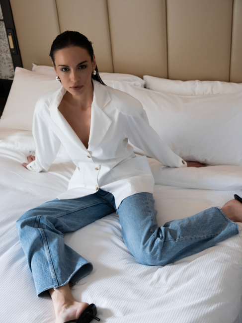 Woman wearing white blazer and blue jeans sitting on a bed looking at camera.