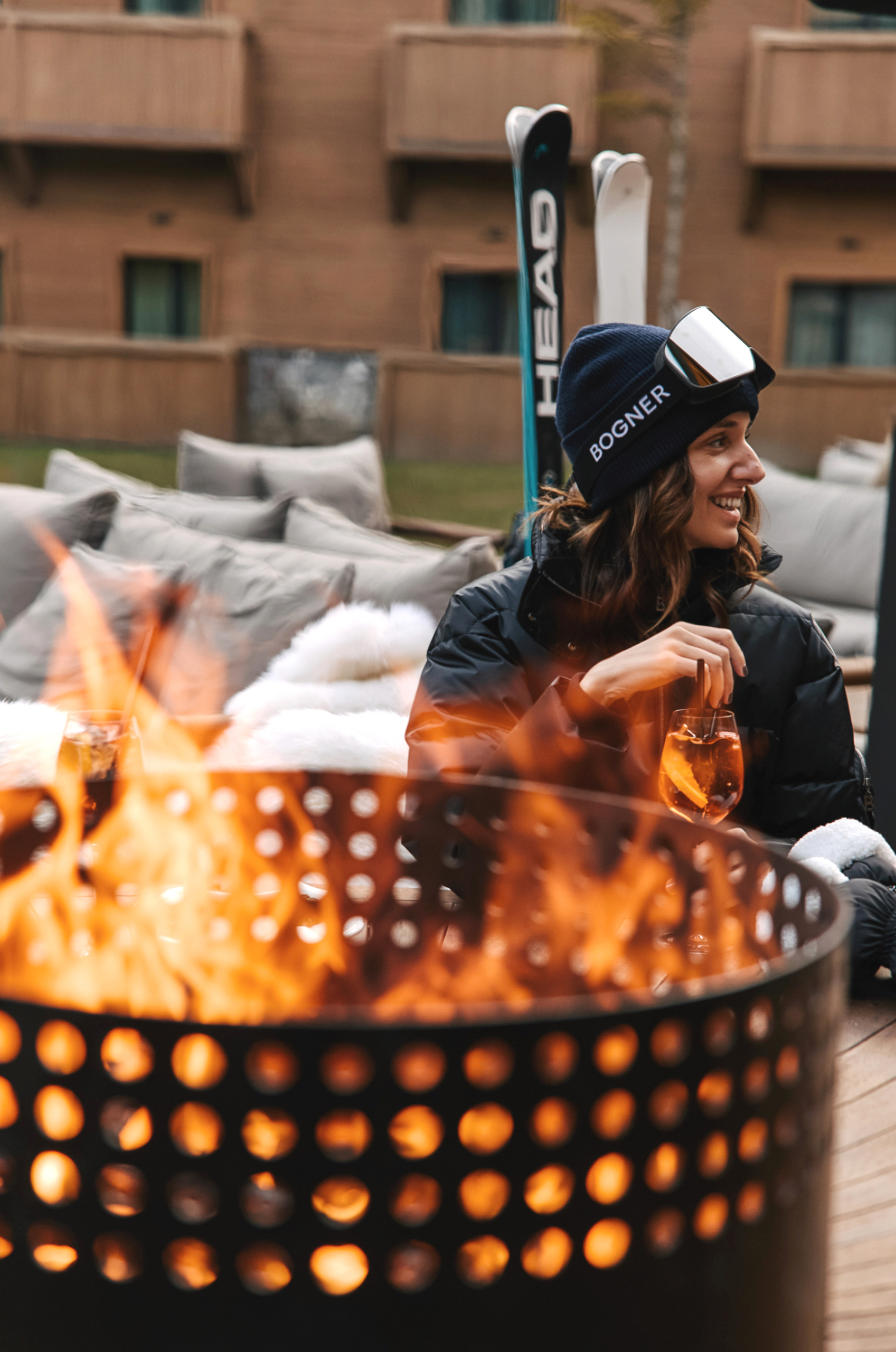 Woman smiles by fire pit with ski gear, drinks, and outdoor seating