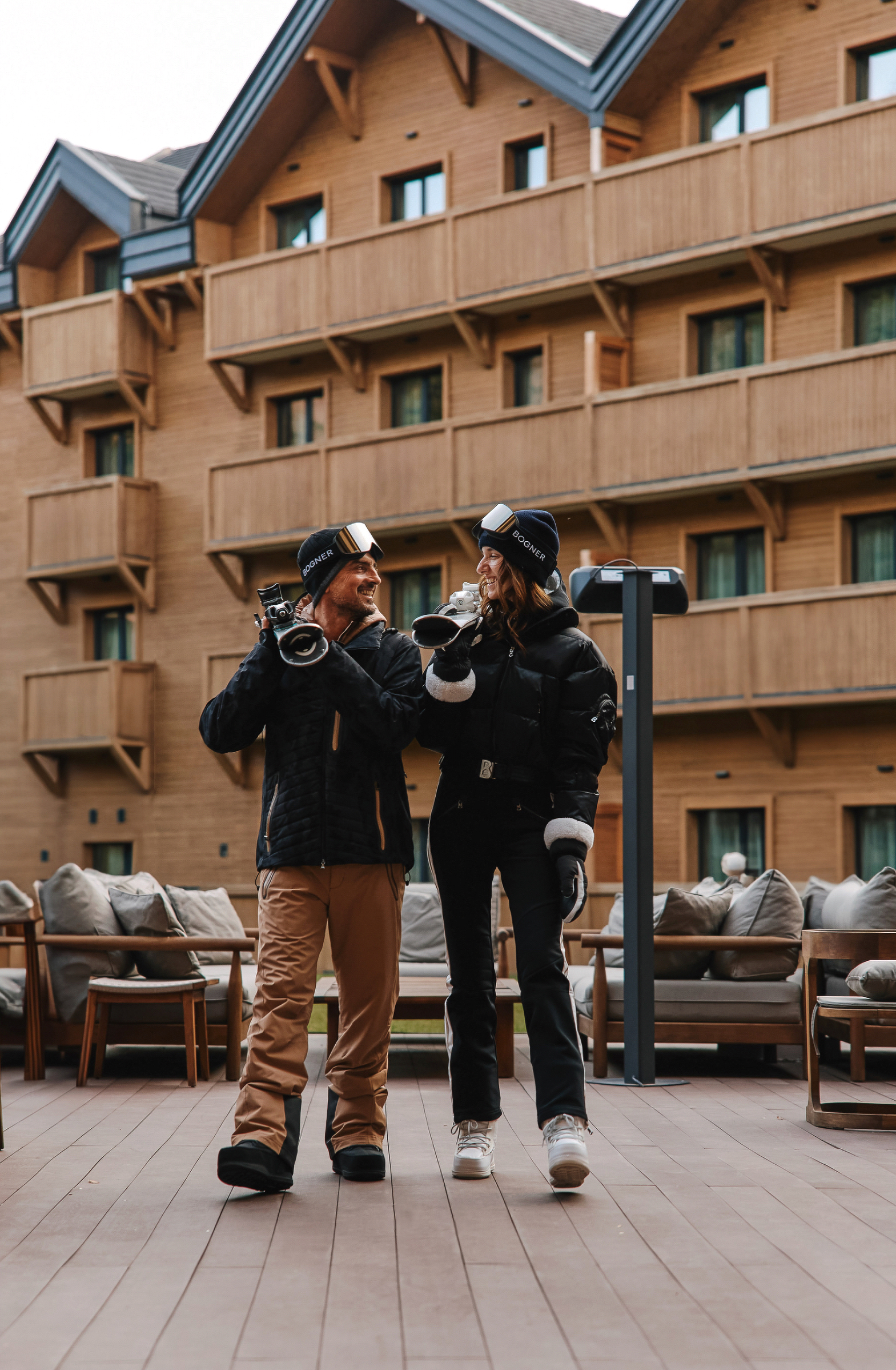 Couple in ski gear smiling while standing on a wooden deck near hotel.