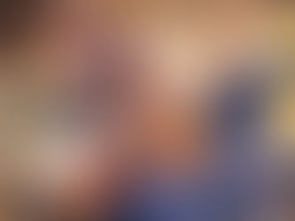 A group of four musicians sit together on the floor in a rehearsal space, thoughtfully engaged with a notebook. One woman in a plaid dress reads from the book while the others—two men with guitars and another with a hat—listen intently. Musical instruments and amplifiers are visible in the background, suggesting a collaborative songwriting session.