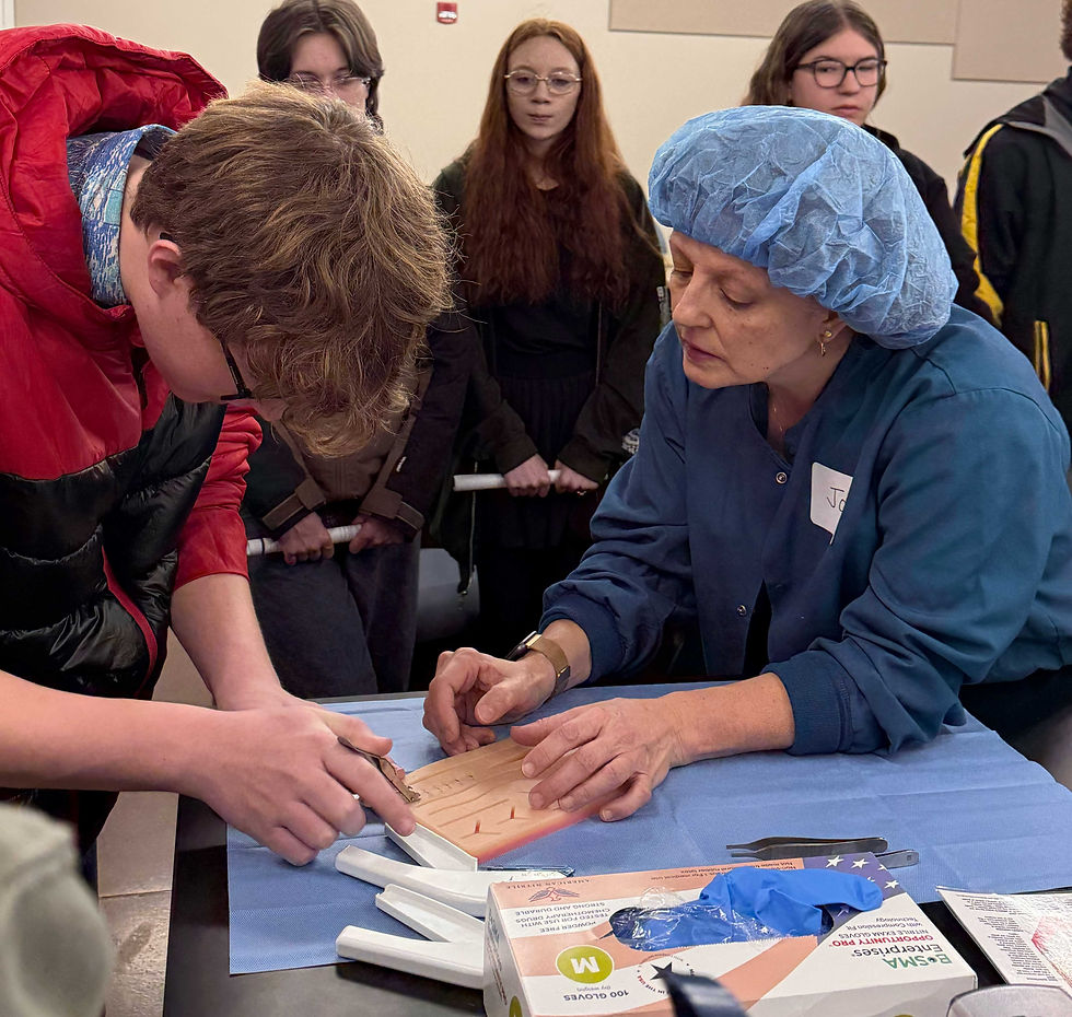 Adult in blue hair cap guides student in red jacket practicing suturing on a pad. Observers watch in classroom setting, focused atmosphere.