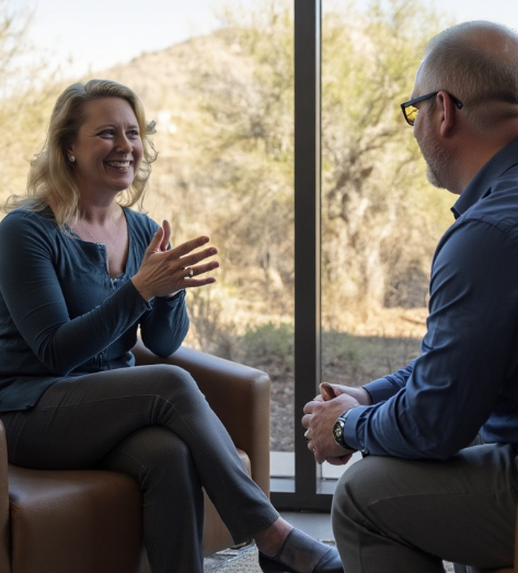 A picture of a women interviewing a man sitting by a window with the sun and trees outside.