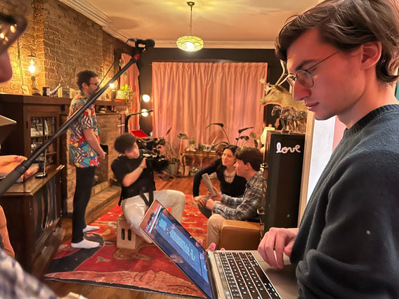 Nathaniel studying a screenplay on  his laptop while a scene is being filmed behind him