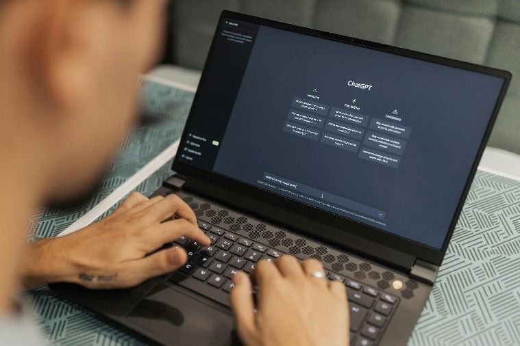 Person typing on a laptop displaying ChatGPT interface. The setting includes a teal-patterned tablecloth. Hands show a tattoo.