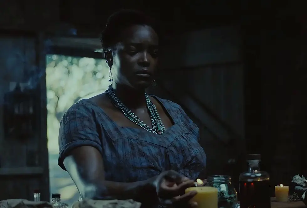 Mulher negra em sala escura acende vela, com colares azuis e expressão séria, diante de garrafas e potes.