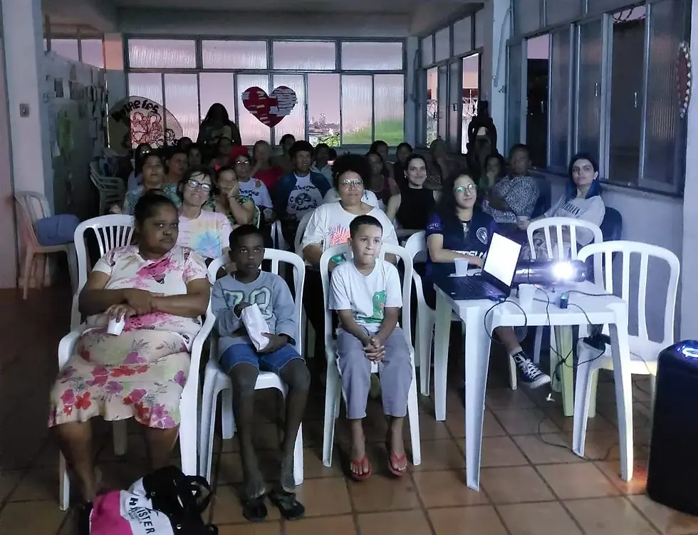 Grupo de adultos e crianças sentados em cadeiras plásticas, assistindo a uma projeção com laptop e projetor em sala iluminada.