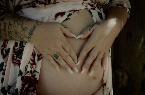 Pregnant woman forms heart over belly, Maternity, floral robe, tattoos are visible.