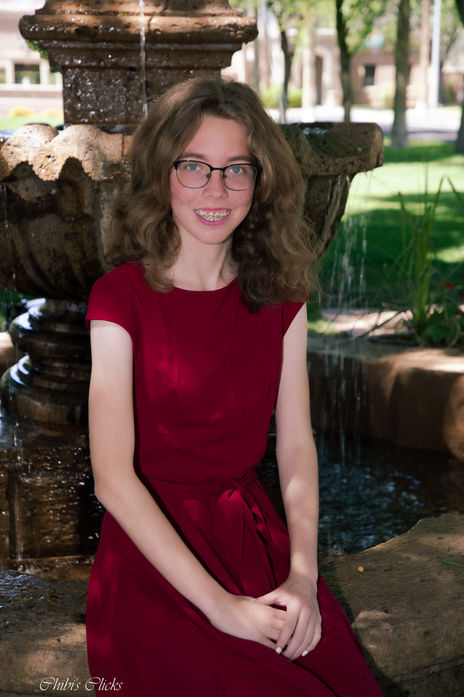 Young woman in red dress smiles near fountain, Senior Portrait, outdoors.