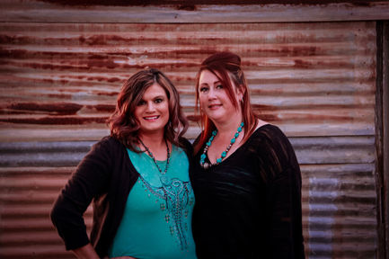 Two smiling women posing, turquoise necklace, black sweater, background of rusty metal.