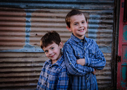 Two boys smiling, wearing blue shirts, standing beside corrugated metal wall.