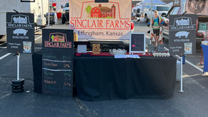 The Sinclair Farms booth with black signs with products listed and a table with black tablecloth sit under a blue pop up tent 