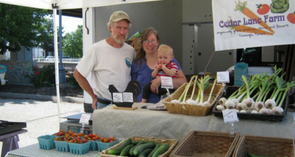 A man and a woman holding a baby stand behind tables full of produce