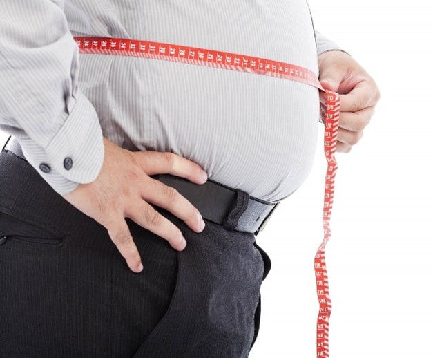 A person measures their waist with a red measuring tape, wearing a gray dress shirt and black pants, suggesting concern about size.