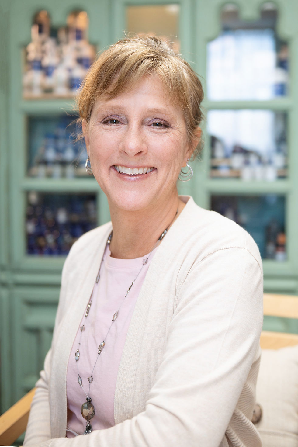 Smiling woman in a light sweater and necklace, seated in front of a teal cabinet with blurred jars, conveying a cheerful mood.