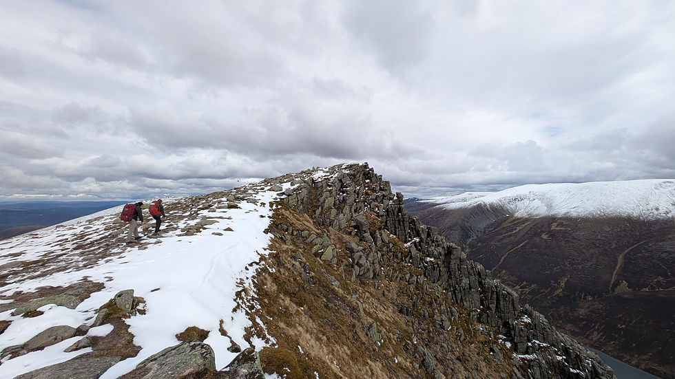 Two hikers with red backpacks trek snowy mountain ridge under cloudy sky, overlooking expansive winter landscape. Rugged, adventurous scene.