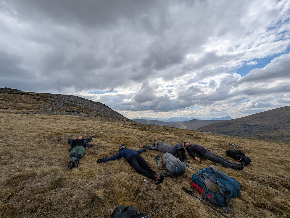 People lying on grassy hillside under cloudy sky, backpacks and hiking poles nearby. Relaxing mood with distant mountains in background.