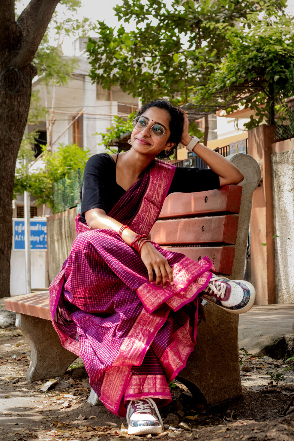 Divya is seated on a bench, one arm resting on the back of it with her head supported by this hand, the other arm is in her lap. She has one leg crossed over the other and is wearing a pink saree with white sneakers. Taken during her outdoors / street portrait session.