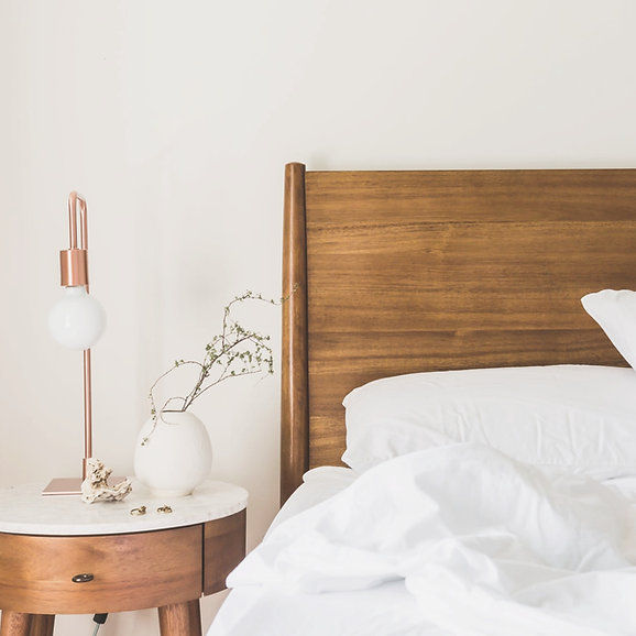 Bright bedroom with a wooden headboard, white bedding, and a round side table holding a copper lamp and white vase, calm and minimal.
