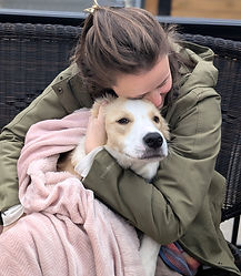 A young women in a green jacket hugging a beige and white dog. He is wrapped in a pink blanket