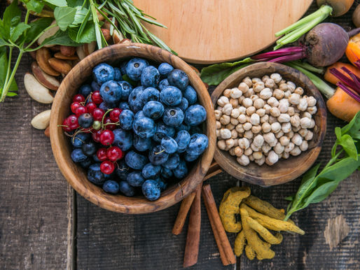 Frutas e grãos em tigelas de madeira sobre mesa rústica, com mirtilos, groselhas, grão-de-bico, ervas e legumes coloridos.