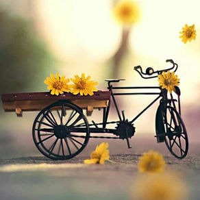 A miniature black tricycle with a wooden cart holds yellow flowers, set against a blurred background with warm tones and floating blooms.