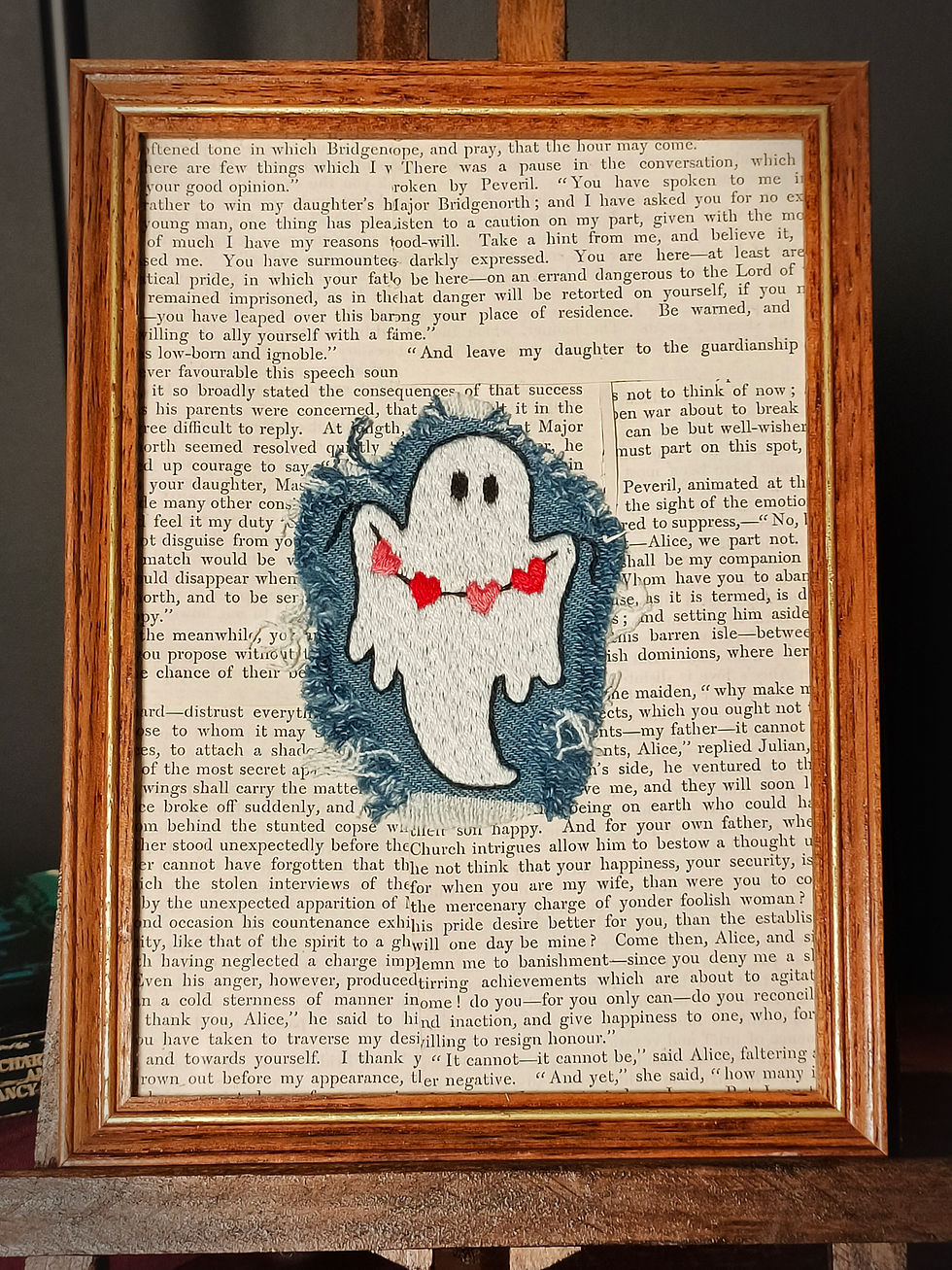 wooden frame, old book page, frayed denim patch, white embroidered ghost holds alternating pink and red heart bunting
