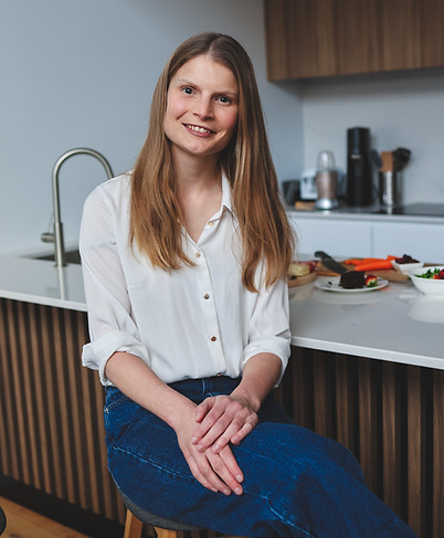 Blonde lady sitting on a stool in a kitchen smiling warmly.