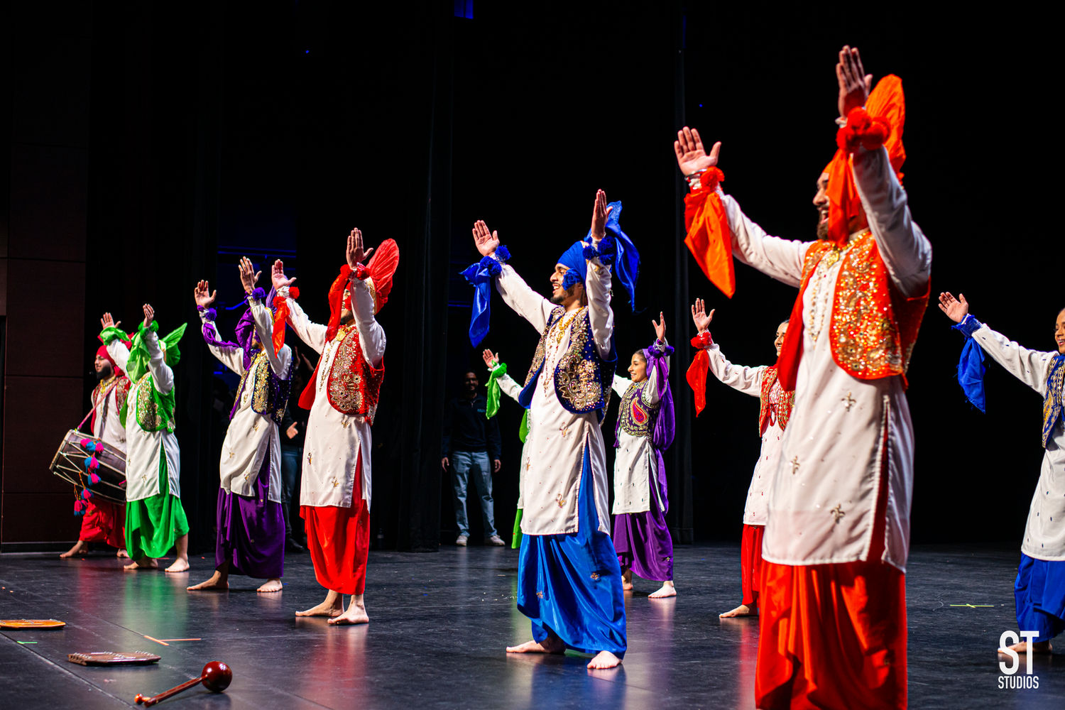 Bhangra dancers throw both arms up as the dholi plays at the back of the stage.